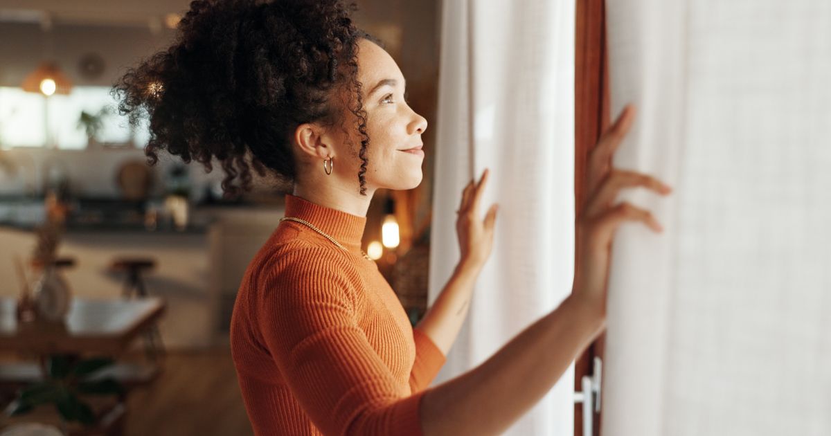 Young woman smiling while opening the curtains in her home.